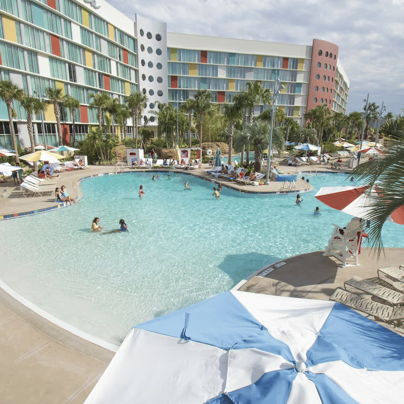 🏖️ Cabana Bay Beach Resort Review with Volcano Bay View – A Fantastic Family Hotel 5 Large resort pool with people swimming, surrounded by palm trees and a colourful retro-style hotel. Cabana Bay Beach Resort Review with Volcano Bay View