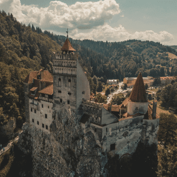 Compare Flights and Travel Essentials 32 Aerial view of Bran Castle in Romania, perched dramatically on a rocky hilltop surrounded by lush green forests, under a partly cloudy summer sky.