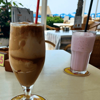 Ayia Napa in May – A Mum & Daughter Escape 🌊☀️ 29 Glass of iced coffee and strawberry milkshake on a seaside café table with beach umbrellas in the background