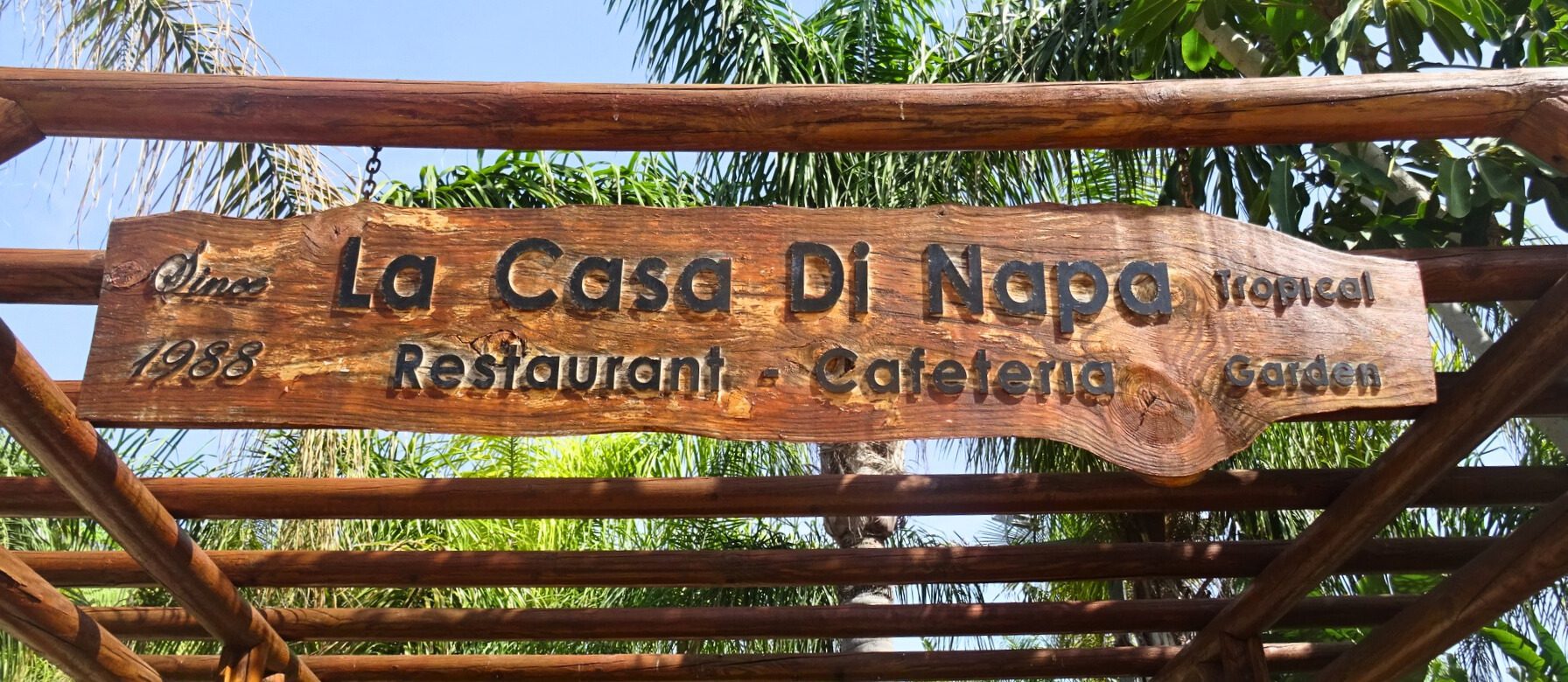 Ayia Napa in May – A Mum & Daughter Escape 🌊☀️ 10 Wooden sign for La Casa Di Napa tropical restaurant and cafeteria, surrounded by lush palm trees.