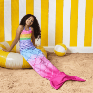 Smiling girl sitting on an inflatable ring wearing a colourful mermaid tail towel against a yellow and white striped background. OHS kids wearable towel