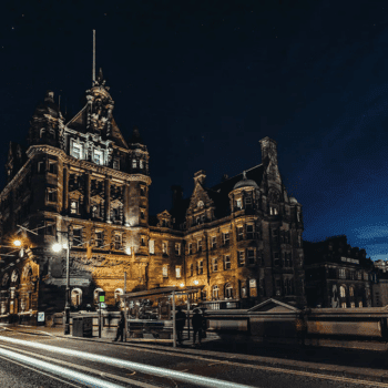 🎃 Spooky Season Travel Deals & Haunted Adventures ✈️👻 10 Night view of The Scotsman Hotel in Edinburgh, illuminated against the dark sky with light trails from passing traffic