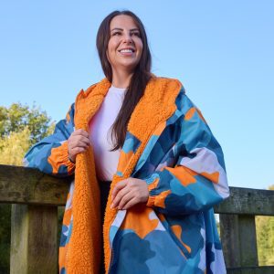 Compare Flights and Travel Essentials 54 Woman wearing the OHS Adults Water Resistant Camo Sherpa Drying Robe – Blue/Orange by a lakeside, showing cosy sherpa lining and long protective fit