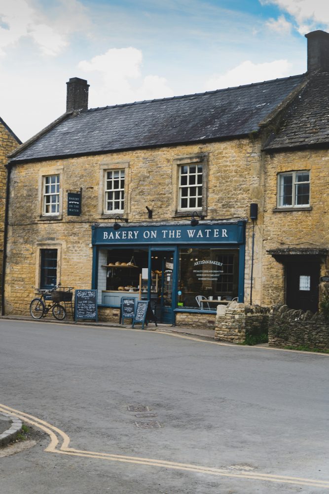Compare Flights and Travel Essentials 80 Bakery on the Water stone building in Bourton-on-the-Water village, Cotswolds