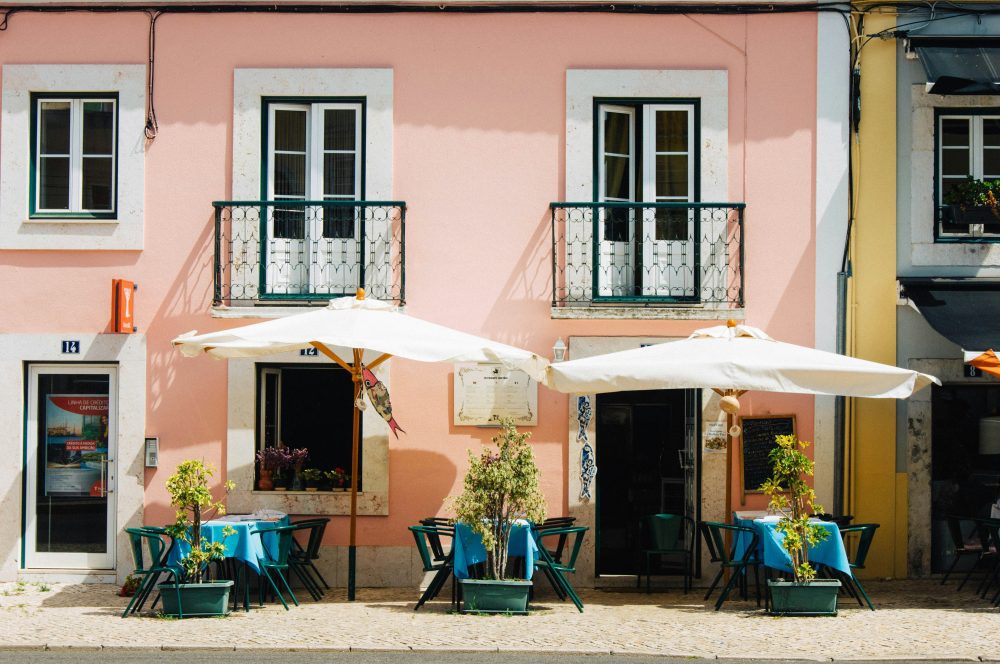 Compare Flights and Travel Essentials 100 Pastel-coloured buildings and outdoor café tables on a quiet Lisbon street