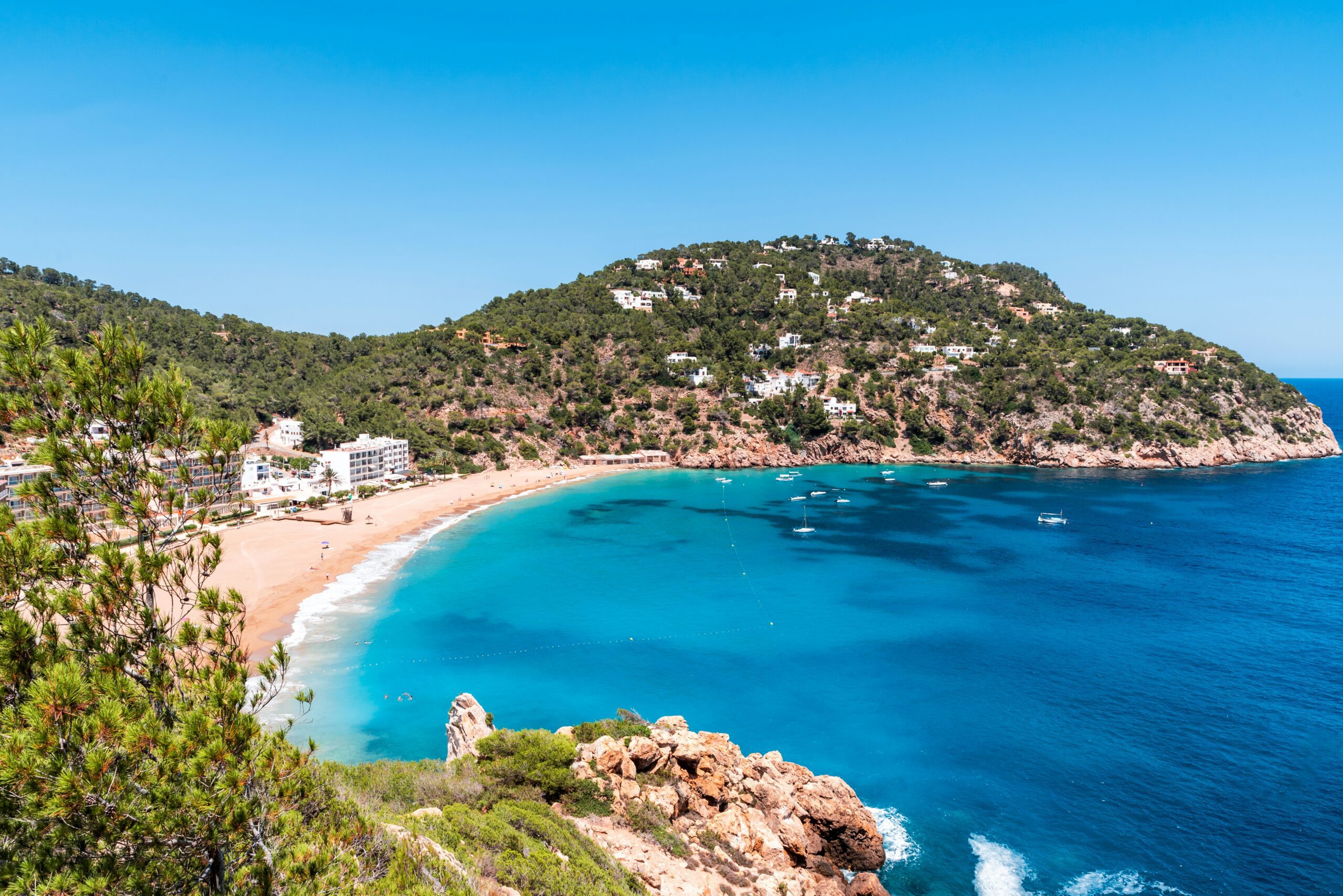 Clear turquoise bay with sandy beach, boats on calm water and hillside villas overlooking the coast