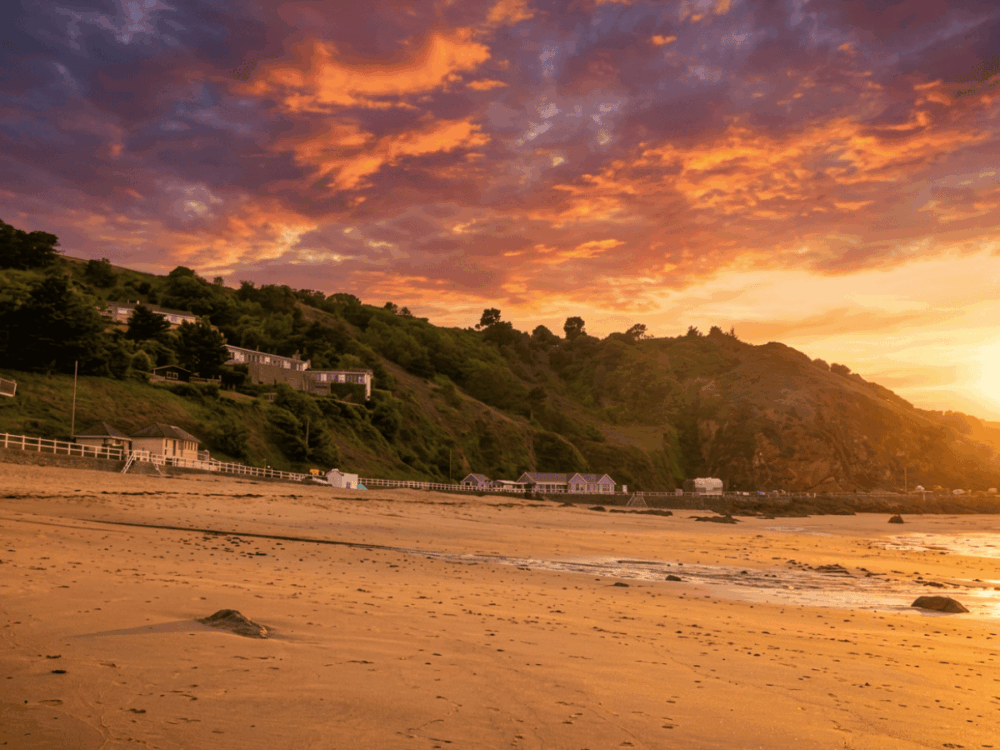 Wide sandy beach with hillside homes under a colourful sunset sky.