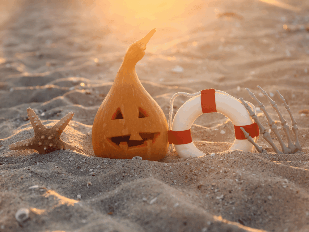 Halloween pumpkin on a sandy beach with a lifebuoy and skeleton hand at sunset
