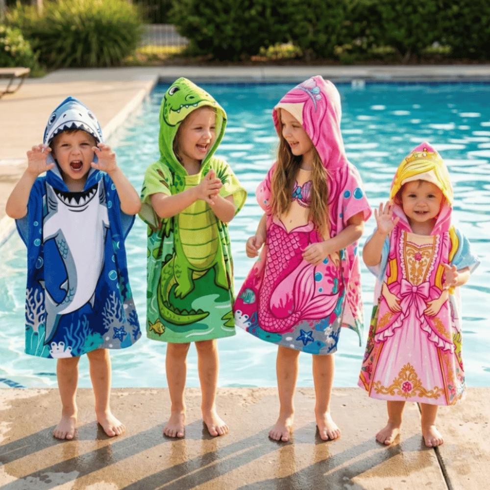 Four children wearing blue shark, green crocodile, pink mermaid and pink princess hooded poncho towels beside a swimming pool.