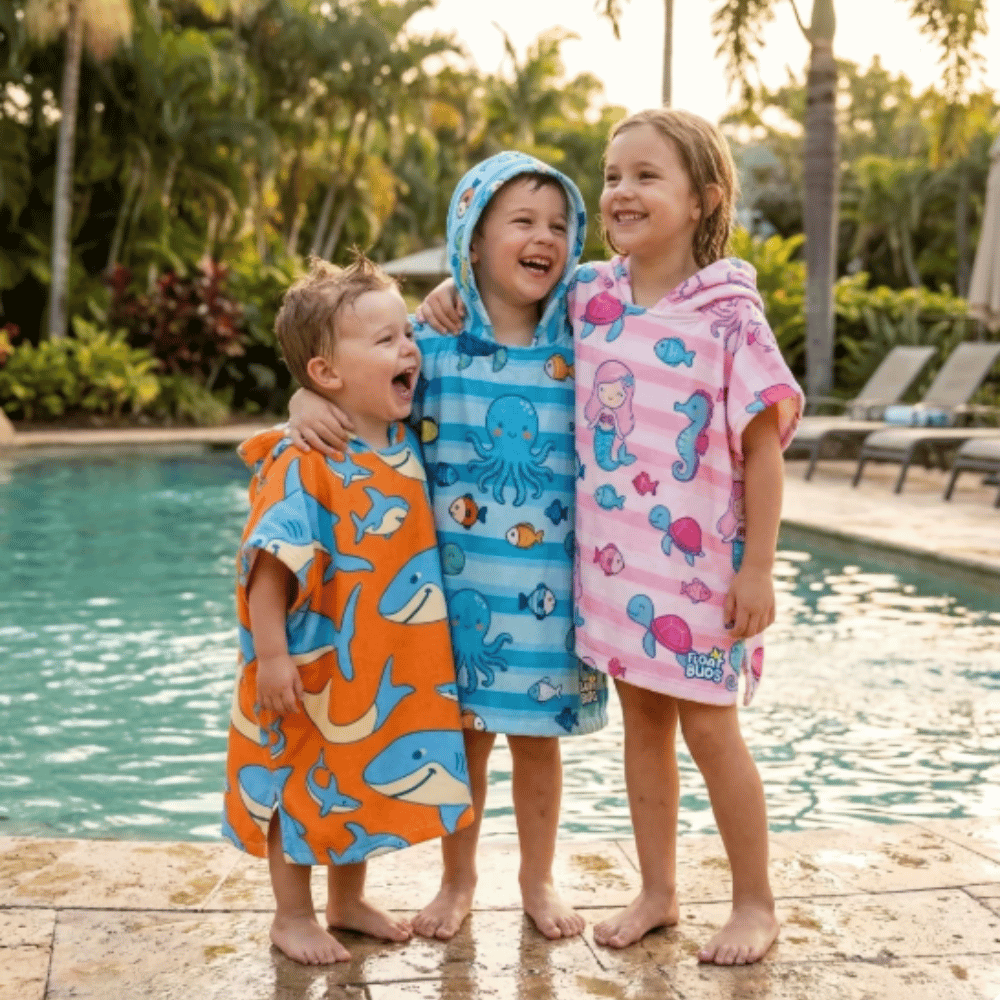 Three children wearing orange shark, blue sea-life and pink mermaid FloatBuds hooded poncho towels beside a swimming pool.