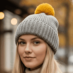 Woman wearing a light grey knitted beanie with a folded ribbed brim and mustard pom pom.