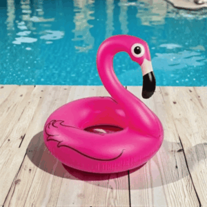 Bright pink inflatable flamingo pool float standing on a wooden poolside deck with blue swimming pool water in the background.