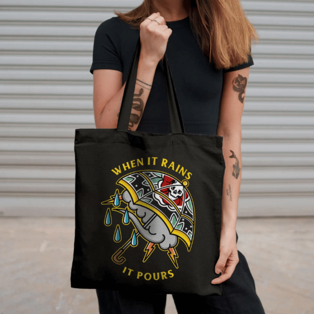 Black tote bag with long handles and colourful When It Rains It Pours umbrella artwork, carried over the shoulder by a person wearing black.
