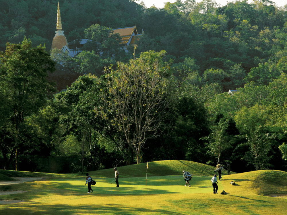 Golfers on a green beneath a wooded hillside and golden temple spire in Hua Hin.