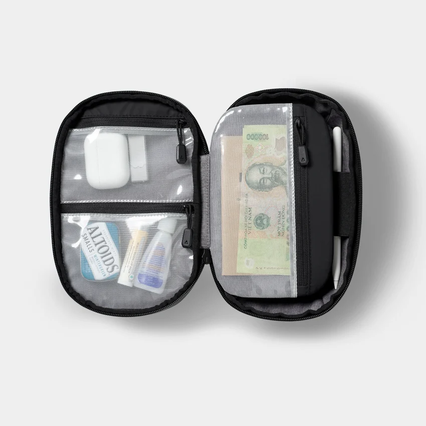 Open black Nomatic Navigator Tech Organizer showing mesh zippered pockets containing small accessories and banknotes, with a white pen secured at the side.