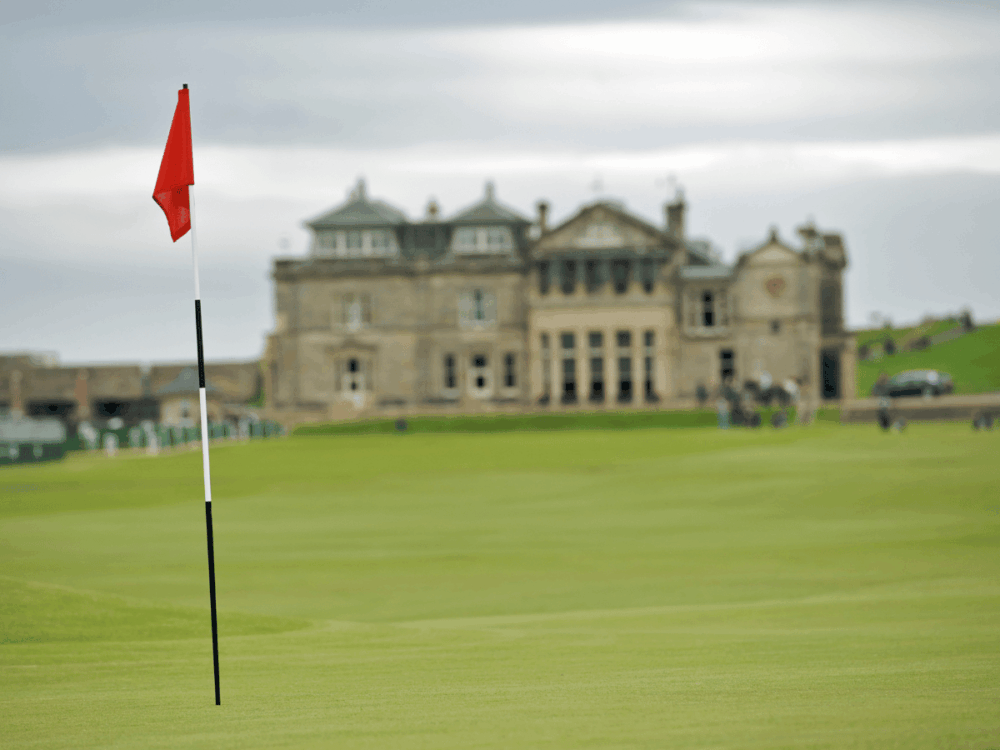 Red golf flag on a green with a large stone building behind in St Andrews, Scotland.